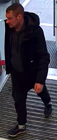 Mężczyzna w wieku około 30-35 lat, ubrany w czarną kurtkę z kapturem, ciemne spodnie jeansowe oraz sportowe buty