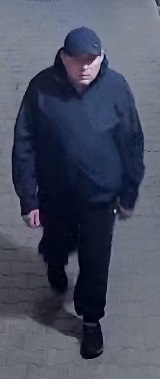 mężczyzna w wieku około 40 lat, krępej budowy ciała, ubrany w czarną kurtkę z kapturem, czarną czapką z daszkiem, czarne spodnie, oraz ciemne buty