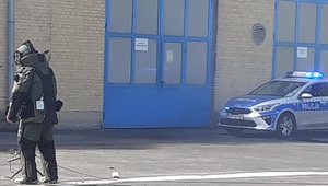 Radiowóz policyjny, a przed nim pirotechnik badający podejrzany ładunek.
