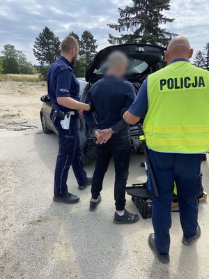 Dwóch policjantów trzyma mężczyznę za ręce, który skuty jest kajdankami.