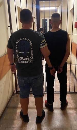 Nieumundurowany funkcjonariusz z opaską na ramieniu z napisem policja prowadzi zatrzymanego.