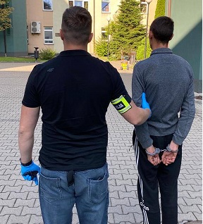 Policjant trzyma zatrzymanego mężczyznę, który ma kajdanki założone na ręce trzymane z tyłu.