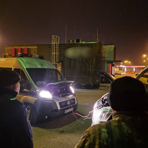 Policjant i właściciel uruchamianego pojazdu. W tle pojazdy z podłączonymi kablami rozruchowymi.