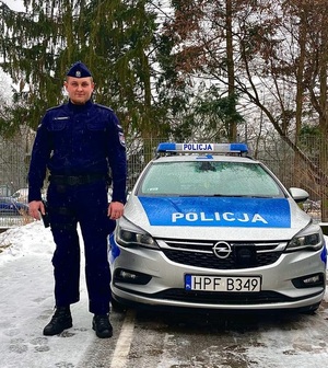 Umundurowany policjant stojący obok oznakowanego radiowozu.