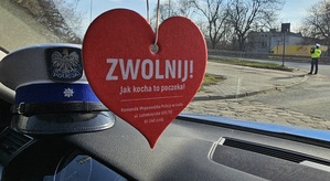 Serduszko z napisem jak kocha to poczeka zawieszone w radiowozie. Na podszybiu czapka policjanta ruchu drogowego. W tle funkcjonariusz ruchu drogowego w trakcie pomiaru prędkości.