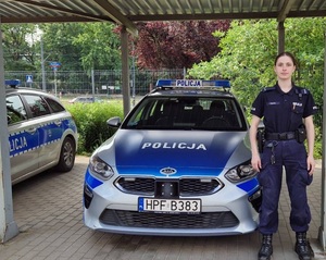 Umundurowana policjantka stoi koło oznakowanego radiowozu policyjnego.
