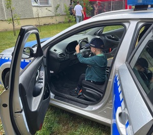 Chłopiec w czapce policyjnej za kierownicą oznakowanego radiowozu.