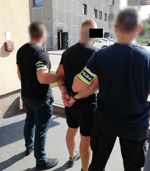 Policjanci trzymają mężczyznę, który ma kajdanki na ręce trzymane z tyłu.
