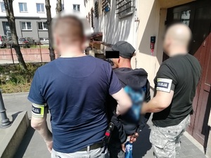 Policjanci trzymają mężczyznę, który ma kajdanki założone na ręce trzymane z tyłu.