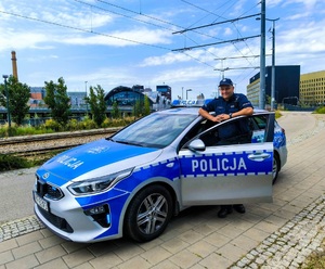 Umundurowany policjant stoi przy oznakowanym radiowozie.