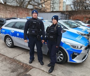 umundurowani policjantka i policjant stoją na tle oznakowanych radiowozów.