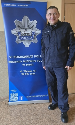 Umundurowany policjant na tle roll-up VI Komisariatu Policji Komendy Miejskiej Policji w Łodzi.