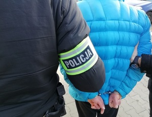 Policjanci trzymają zatrzymanego mężczyznę, który ma założone kajdanki na ręce z tyłu.