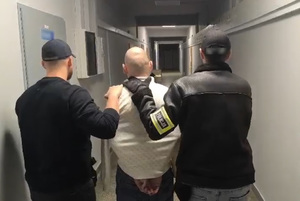 Policjanci prowadzą zatrzymanego mężczyznę, który ma założone kajdanki na ręce z tyłu.