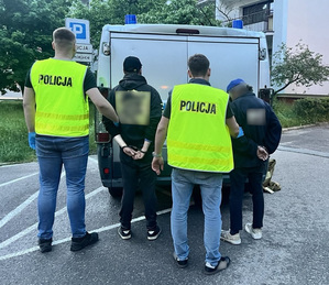 Policjanci trzymają zatrzymanego mężczyznę przed radiowozem.