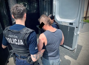Umundurowani policjanci pomagają zatrzymanemu mężczyźnie wsiąść do radiowozu.
