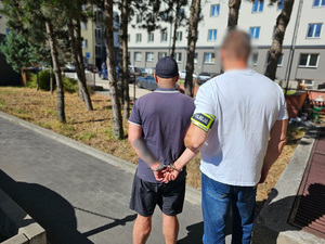 Policjant trzyma mężczyznę, który ma kajdanki na ręce trzymane z tyłu.