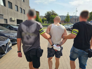 Policjanci prowadzą mężczyznę, który ma założone kajdanki na ręce z tyłu.