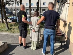 Policjanci trzymają kobietę, która ma kajdanki założone na ręce z tyłu.