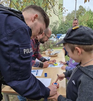 Chłopiec przy umundurowanym policjancie robi odcisk linii papilarnych na kartce.