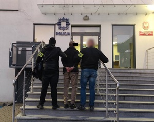 Policjanci stoją z zatrzymanym mężczyzną na schodach przed komisariatem.