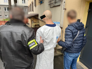 Policjanci trzymają mężczyznę, który ma założone kajdanki zespolone.