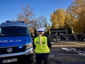 Policjantka stoi obok radiowozu na przeciwko cmentarza.