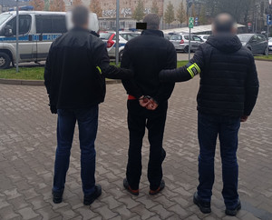 Policjanci trzymają mężczyznę, który ma kajdanki założone na ręce z tyłu.