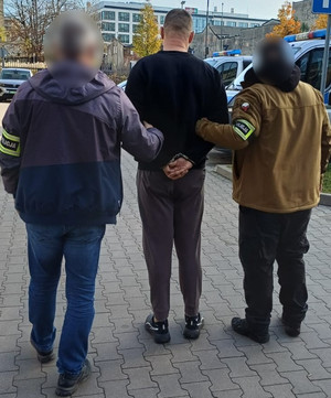 Policjanci prowadzą mężczyznę, który ma kajdanki założone na ręce z tyłu.