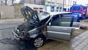 Spalone auto, w tle strażacy w trakcie akcji i wóz gaśniczy.