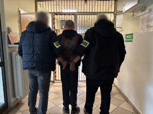 Policjanci prowadzą mężczyznę, który ma założone kajdanki na ręce z tyłu.