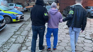 Policjanci prowadzą mężczyznę, który a założone kajdanki zespolone.