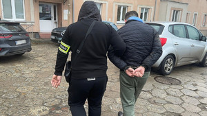 Policjant prowadzi mężczyznę, który ma założone kajdanki na ręce z tyłu.