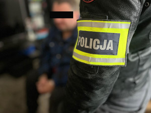 Opaska z napisem Policja na ramieniu policjanta, w tle siedzi mężczyzna.