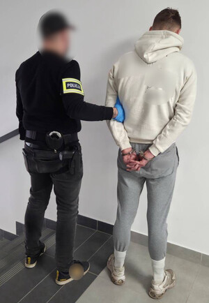 Policjant trzyma mężczyznę, który ma założone kajdanki na ręce z tyłu.