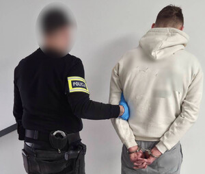 Policjant trzyma mężczyznę, który ma założone kajdanki na ręce z tyłu.