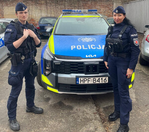 Umundurowana policjantka i policjant stoją przed oznakowanym radiowozem.