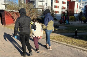 Policjanci prowadzą kobietę, która ma założone kajdanki na ręce z tyłu.