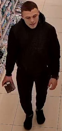 Mężczyzna ubrany w czarną kurtkę na długi rękaw, czarne spodnie dresowe, szare skarpetki i czarne buty typu sportowego. Średniego wzrostu, średniej budowy ciała, z czarnymi krótko ściętymi włosami i lekko odstającymi uszami.