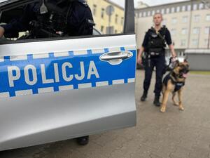 Drzwi radiowozu z napisem policja, w tle policjantka z psem służbowym.