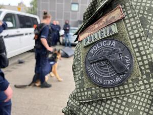 Naszywka policyjnego instruktora wyszkolenia strzeleckiego na rękawie munduru, w tle policjanci w trakcie szkolenia.