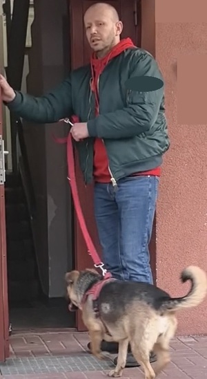 Pies razem z opiekunem stoją przed drzwiami wejściowymi do bloku.