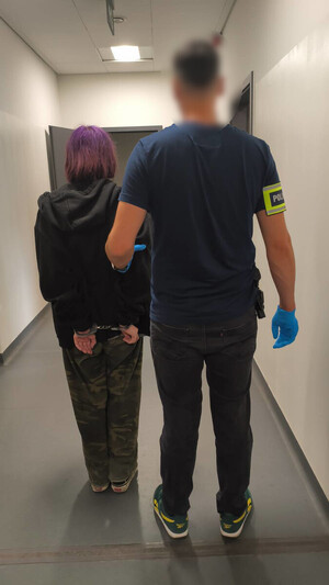 Policjant trzyma kobietę, która ma założone kajdanki na ręce z tyłu.