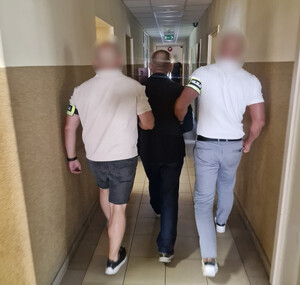 Policjanci prowadzą mężczyznę, który ma założone kajdanki na ręce z tyłu.