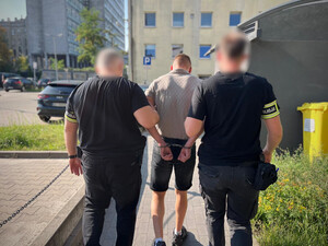 Policjanci prowadzą mężczyznę, który ma kajdanki założone na ręce z tyłu.