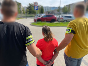 Policjanci trzymają kobietę, która ma założone kajdanki na ręce trzymane z tyłu.