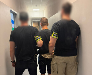 Policjanci prowadzą mężczyznę, który ma założone kajdanki na ręce z tyłu.