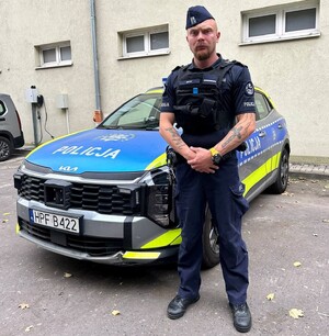 Umundurowany policjant stoi przed oznakowanym policyjnym radiowozem.