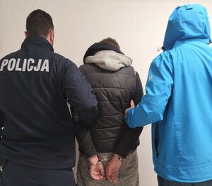 Policjanci trzymają mężczyznę, który ma założone kajdanki na ręce trzymane z tyłu.
