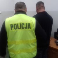 Mężczyzna stojący obok policjanta ubranego w kamizelkę odblaskową z napisem policja. Mężczyźni stoją tyłem do obiektywu. Policjant prowadzi mężczyznę podtrzymując go za ramię